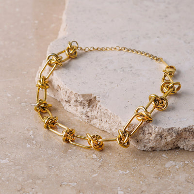  The Valerie Knotted Bracelet is showcased on a beige, textured stone surface. The gold-toned bracelet features a series of elongated links connected by intricate, polished knots, creating a bold and sculptural design.