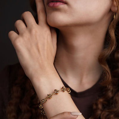 The Valerie Knotted Bracelet is presented in a studio shot on a clean white background, arranged in a circular shape. The gold-toned bracelet is composed of a series of elongated links that are intertwined with smaller knotted links, with a clasp and extender chain visible at the top.