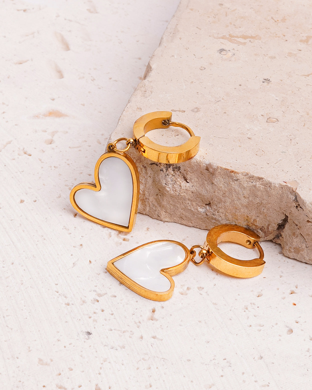 A pair of Venus Heart Drops earrings are showcased on a beige, textured stone surface. The gold-toned hoops are connected to polished heart-shaped charms with a bright, iridescent white inlay.