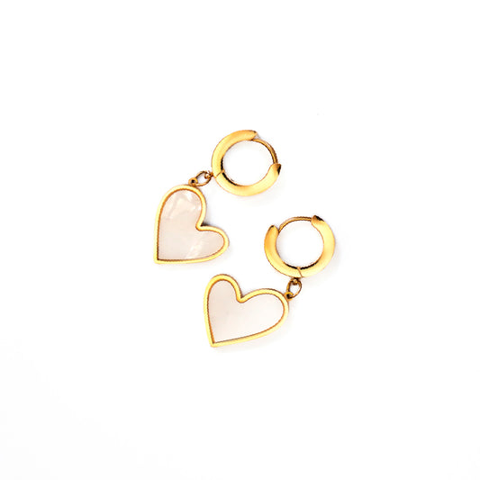 A top-down studio shot against a white background displays a pair of Venus Heart Drops earrings. Each earring consists of a small, polished gold-toned hoop from which a heart-shaped charm, featuring a pearlescent white inlay bordered by gold, dangles.