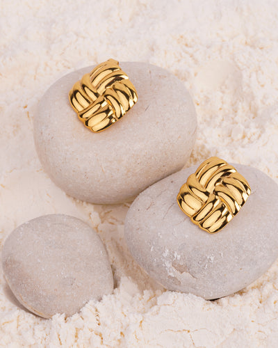 The Venus Knot Studs are showcased on two smooth white stones in a bed of white powder. The gold-toned square stud earrings feature an intricate, raised woven or knot-like pattern.