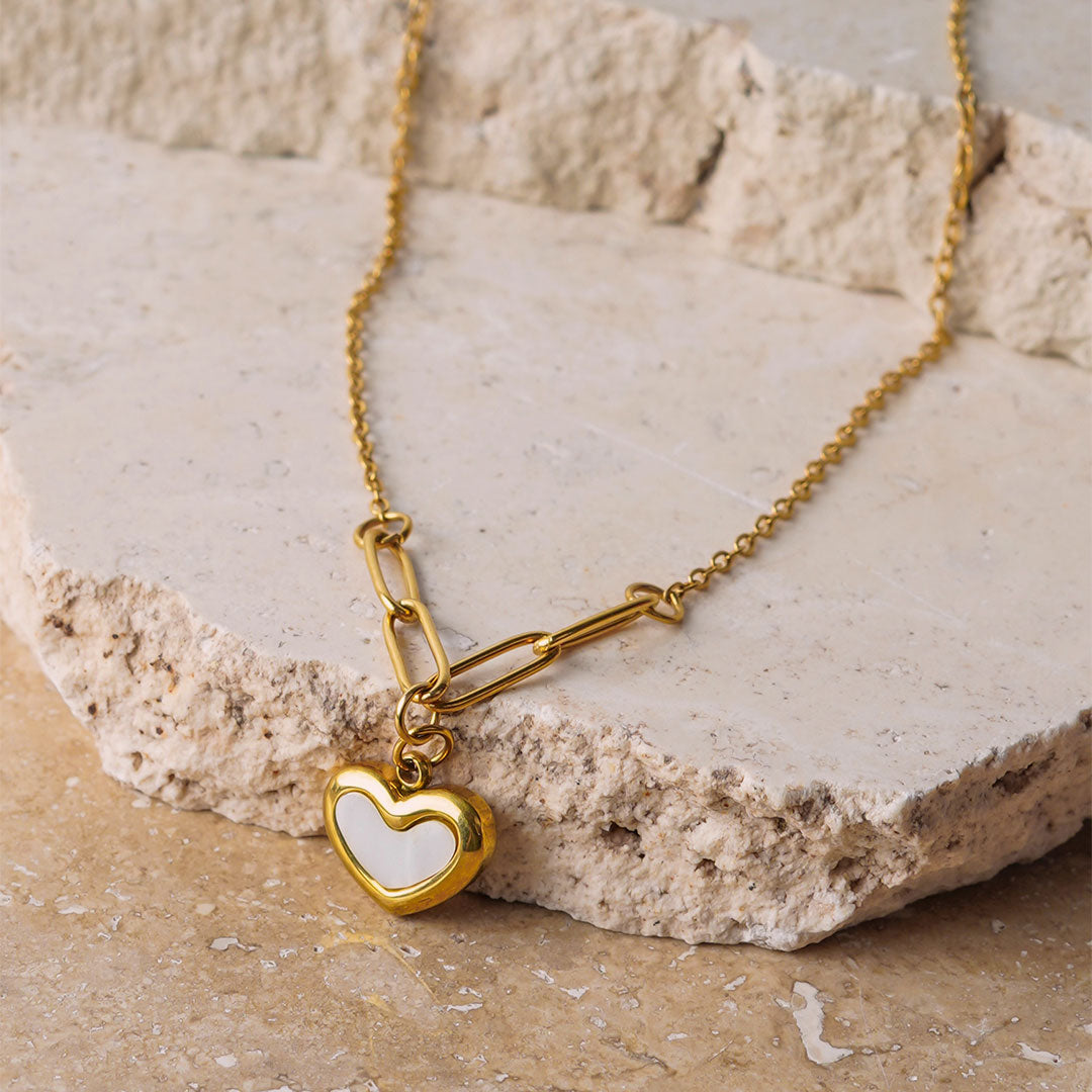 The Violet Petal Pendant is displayed on a textured, beige stone surface. This gold-toned necklace has a unique chain of both fine and elongated links that connect to a striking heart pendant, which has a white, enamel-like insert.