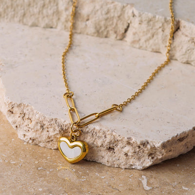The Violet Petal Pendant is displayed on a textured, beige stone surface. This gold-toned necklace has a unique chain of both fine and elongated links that connect to a striking heart pendant, which has a white, enamel-like insert.