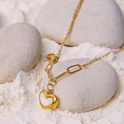 The Violet Petal Pendant rests on a smooth, large white stone surrounded by other stones and white powder. The necklace features a two-part chain (delicate and paperclip links) leading to a heart-shaped pendant with a shiny white center.