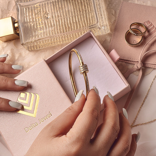 Hands gently opening a Delta Jewel pink box, revealing an exquisite 18k gold plated Elle Minimal Bracelet. Nearby, an 18k gold plated Rose Bloom Ring, a perfume bottle, and a branded pouch complete the luxurious presentation.