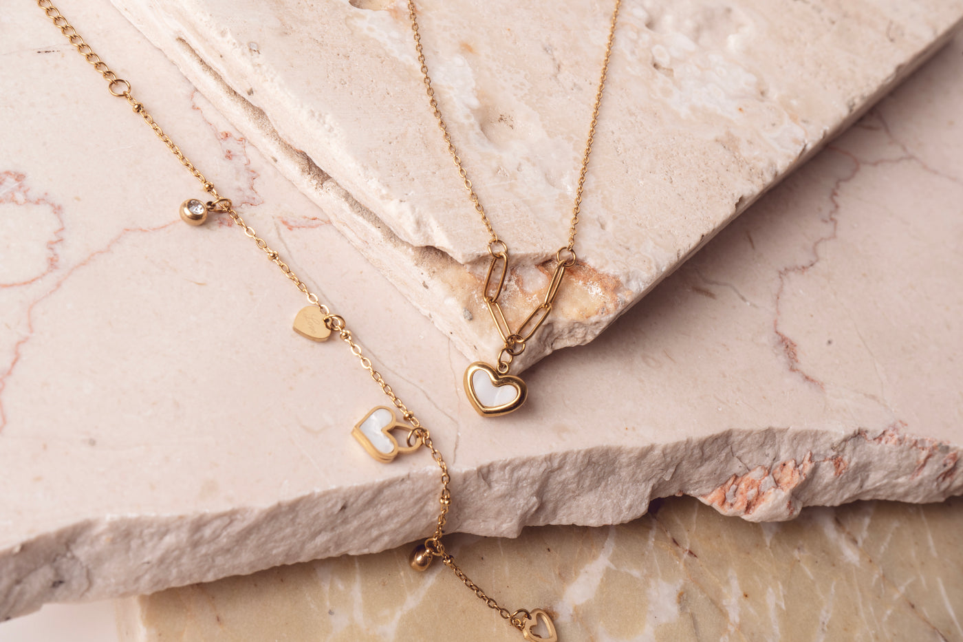 Two delicate 18k gold plated necklaces arranged on a light-colored marble surface. The top necklace is a Violet Petal Pendant, and the bottom necklace is multiple Amara Love Charms, including heart-shaped and sparkling designs by Delta Jewels.
