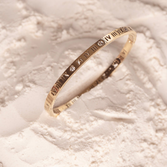 A studio shot on a pristine white background displays the Aria Luxe Bracelet. This polished gold bangle, engraved with Roman numerals, appears to float, with four tiny, clear gemstones shimmering along its surface.