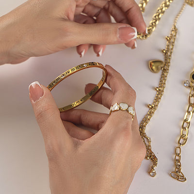 Hands delicately holding an 18k gold plated Ava Grace Bangle adorned with Roman numerals and sparkling accents. On her fingers, an 18k gold plated Hazel Spark Ring and Dahlia Charm Ring are visible, while an Aurora Grace Bracelet, Aria Luxe Bracelet, and Ivy Clover Bracelet are elegantly draped nearby, showcasing a luxurious collection.