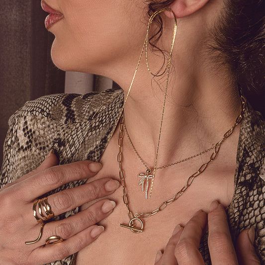 A close-up of a person adorned with a layered collection of 18k gold plated jewelry. This includes an Aria Luxe Bracelet and an Aurora Grace Bracelet (partially visible), a Chloe Forever Necklace and an Elara Grace Pendant adorning the neck, and an 18k gold plated Lila Eternity Band and Mila Zircon Ring on the fingers, all complementing a stylish snake print blazer.
