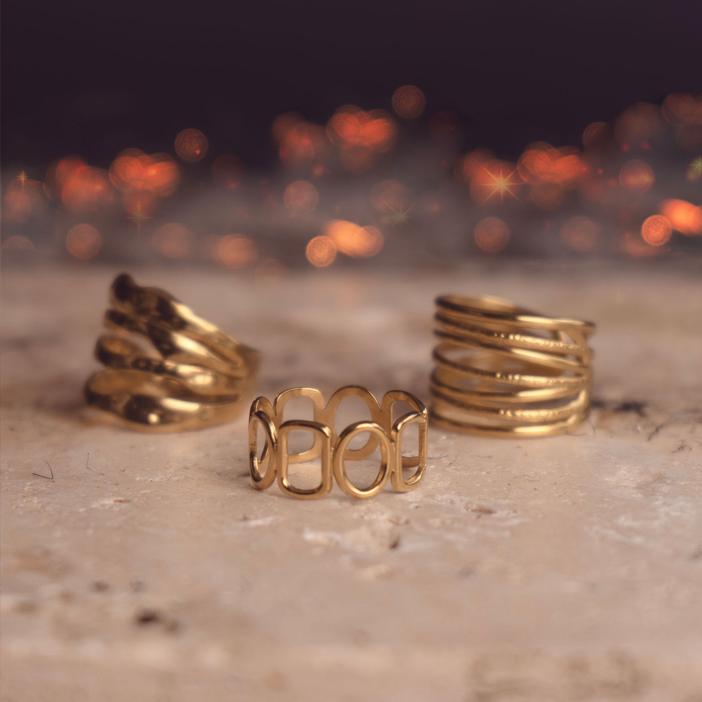 A set of 3 rings including Celeste Radiance Band, Amelia Radiant Ring and Mila Zircon Ring by Delta Jewel. These are modern openwork gold-plated rings, displayed alongside one another on a stone surface with a softly glowing background.