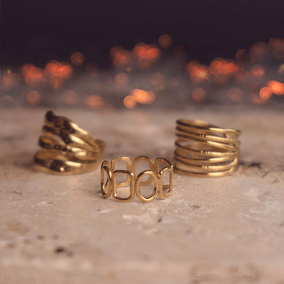 A set of 3 rings including Celeste Radiance Band, Amelia Radiant Ring and Mila Zircon Ring by Delta Jewel. These are modern openwork gold-plated rings, displayed alongside one another on a stone surface with a softly glowing background.